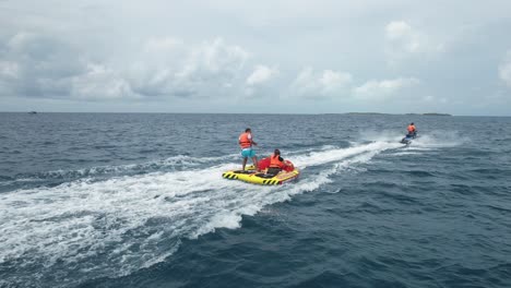 Holidaymaker-Couple-enjoying-Thrill-Ride-on-Water-Tube-Towed-to-Speeding-Jet-Ski,-Drone-Tracking