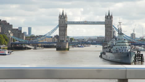 Toma-Dinámica-En-Camión-Del-Tower-Bridge-De-Londres,-Con-El-HMS-Belfast-En-Primer-Plano,-Con-El-Telón-De-Fondo-De-Un-Cielo-Nublado-De-Londres-Durante-El-Día.