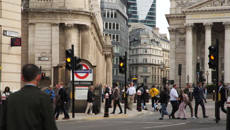 Busy-street-movement-camera-shot-trucking-left-to-underground-Bank-Station,-pedestrians-crossing-at-the-traffic-lights,-with-the-iconic-Bank-of-England-building-towering-in-the-background