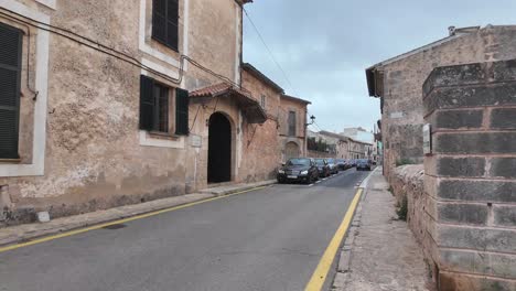 Caminando-Por-La-Calle-En-La-Ciudad-De-Algaida,-Isla-Balear-De-Mallorca-Con-Casas-De-Piedra-Caliza,-Autos-Estacionados-En-La-Calle-En-Abril