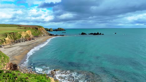 Drone-flying-over-sea-cliffs-people-playing-with-dogs-in-water-dramatic-colours-of-sea-and-skyline-and-rare-rocks-in-emerald-green-seas