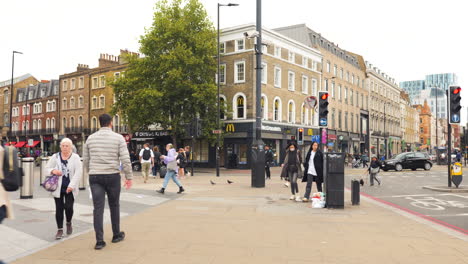 Intersección-Del-Barrio-De-Londres-Repleta-De-Peatones,-En-Medio-De-Múltiples-Movimientos-De-Transporte,-Con-El-Telón-De-Fondo-De-Edificios-A-Escala-Humana-Dentro-De-La-Ciudad.