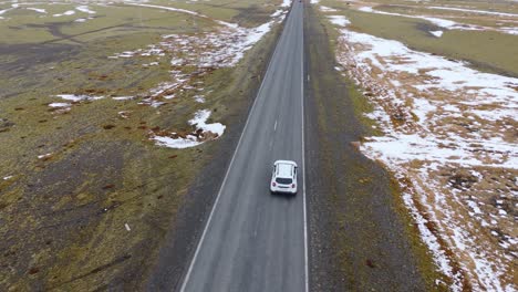 Luftdrohne-Verfolgt-Weißes-Auto-Von-Hinter-Asphaltierter-Straße-Durch-Schneebedeckte-Berge