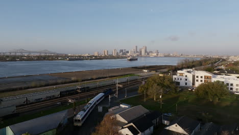 Aerial-flyover-of-a-passenger-train-moving-towards-downtown-with-a-large-container-ship-on-the-Mississippi-River-with-downtown-New-Orleans-in-the-background-in-the-morning