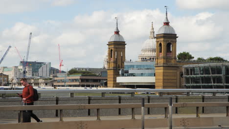 Un-Hombre-Caminando-Por-El-Puente-De-Londres,-Empujando-Una-Maleta-Mientras-Sostiene-Un-Teléfono,-Seguido-Por-Un-Cardán