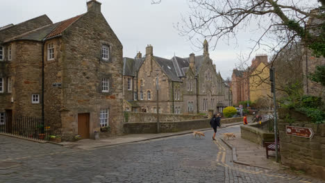 Man-walks-pair-of-dogs-on-cobblestone-road-in-quaint-village-on-outskirts-of-Edinburgh-Scotland