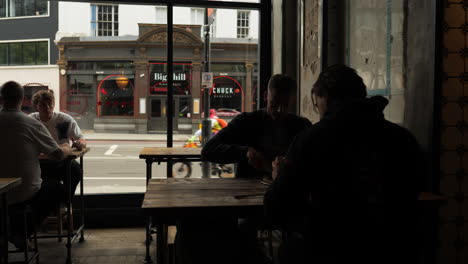 Dimly-lit-restaurant-through-a-static-shot,-as-customers-sit-on-high-chairs-at-tables,-savouring-their-meals-while-observing-the-bustling-cityscape-outside-the-glass-storefront
