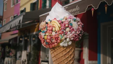 Decorated-ice-cream-cone-in-a-waffle-cone,-with-colorful-flowers,-including-a-pink-rose,-in-the-background-is-a-gelato-shop-with-a-sign-that-says-"Trento"-and-"Elato"-written-in-white-lettering