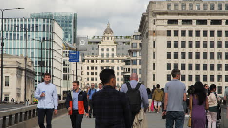 Toma-En-Movimiento-De-Trabajadores-Caminando-Por-El-Puente-De-Londres-Durante-La-Hora-Del-Almuerzo,-Con-Edificios-Emblemáticos-De-La-Ciudad-Al-Fondo