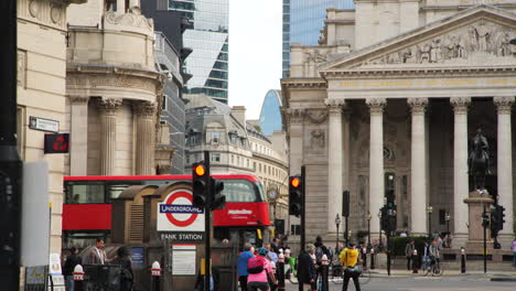 En-La-Bulliciosa-Intersección-Del-Cruce-Bancario,-Los-Peatones-Navegan-Por-Las-Calles-Mientras-El-Icónico-Autobús-Rojo-De-Londres-Pasa-Elegantemente-Al-Fondo.