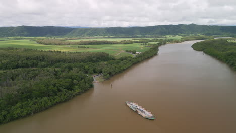 Weiße-Fähre-Fährt-Zwischen-Bewaldeten-Hügeln-Im-Braunen-Flusswasserkanal,-Drohnen-Luftpanorama