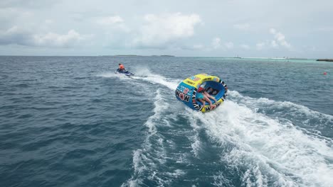 Siga-La-Toma-De-Un-Dron-De-Una-Pareja-Montando-Un-Tubo-De-Esquí-Acuático-Tirado-Por-Una-Moto-Acuática-A-Alta-Velocidad-Contra-La-Costa-De-La-Isla