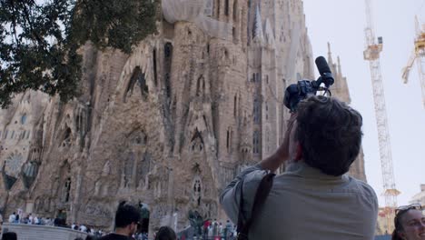 Man-capturing-Sagrada-Familia-on-camera,-Barcelona,-cranes-in-background,-tourists,-daylight