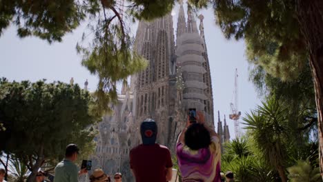 Los-Turistas-Contemplan-La-Sagrada-Familia-Bajo-Un-Cielo-Azul-Brillante,-Enmarcado-Por-Un-Follaje-Verde,-En-Barcelona.