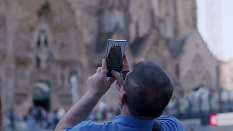 Man-capturing-Sagrada-Familia-with-smartphone,-Barcelona,-tourists-around,-daytime