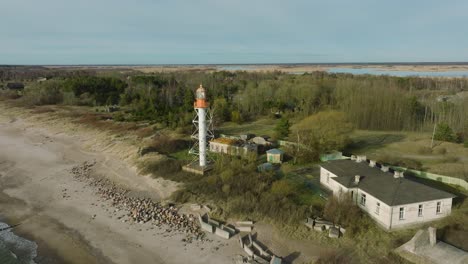 Luftaufnahme-Des-Weißen-Leuchtturms-Von-Pape,-Ostseeküste,-Lettland,-Weißer-Sandstrand,-Große-Wellen-Brechen,-Sonniger-Tag-Mit-Wolken,-Breite-Drohnenaufnahme-In-Vorwärtsbewegung