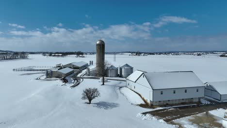 Snowy-farm-with-silo,-barns,-and-outbuildings,-surrounded-by-open-fields-under-a-blue-sky