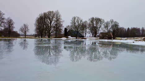 Pan-shot-over-frozen-lake-surrounded-by-wooden-cottages-along-rural-countryside-on-a-cold-winter-day