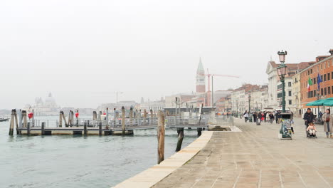 Menschen,-Die-An-Der-Uferpromenade-Entlang-Der-Lagune-Von-Venedig-Spazieren,-In-Der-Ferne-Der-Markusturm-In-Venedig,-Italien