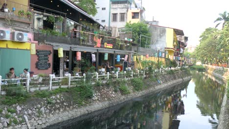 Cafes-lining-a-tranquil-water-channel-in-Hanoi-at-dusk,-locals-dining-al-fresco,-urban-reflection