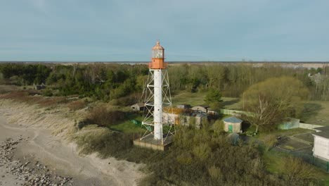 Luftaufnahme-Des-Weiß-Gefärbten-Pape-Leuchtturms,-Ostseeküste,-Lettland,-Weißer-Sandstrand,-Große-Wellen,-Sonniger-Tag-Mit-Wolken,-Weite-Drohnen-Umlaufaufnahme