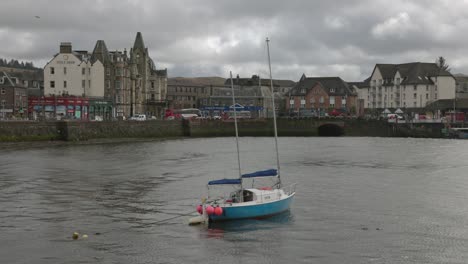 Toma-Estática-De-Un-Pequeño-Barco-Anclado-En-La-Bahía-De-Oban-Con-La-Calle-Principal-Detrás.