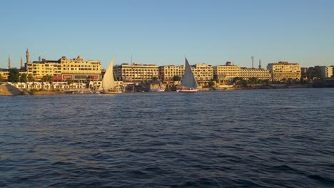 A-sunny-day-in-Egypt-on-the-Nile-River-in-the-city-of-Aswan-features-sailboats,-bustling-riverbanks,-ancient-cityscapes,-and-a-vibrant-city-atmosphere