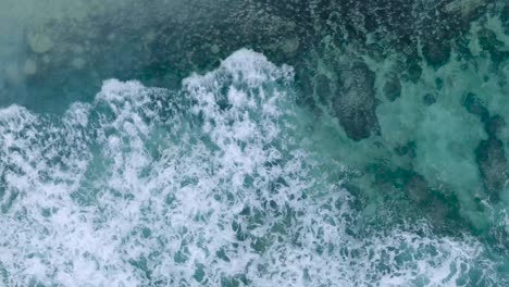 Top-Down-Drone-View-of-turquoise-clear-water-and-crashing-waves-over-shallow-coral-reef-in-Uluwatu-Bali-Indonesia