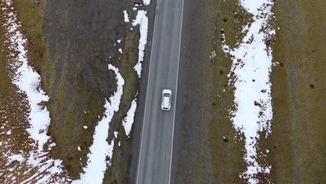 Aerial-Drone-Top-Down-White-Car-Driving-Through-Snowy-winter-road-follow-asphalted-way