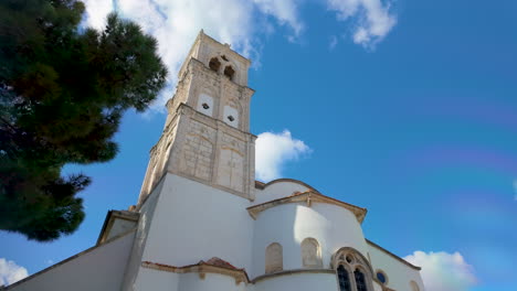 Vista-En-ángulo-Bajo-De-Una-Iglesia-Blanca-Con-Un-Campanario-De-Piedra-Contra-Un-Cielo-Azul-Con-Nubes-Esponjosas,-Rodeada-De-Vegetación,-Ubicada-En-Lefkara,-Chipre