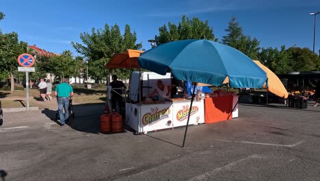 Small-street-stall-preparing-churros,-cakes-and-sweets-with-gas-cylinders-in-the-kitchen-with-oil-under-the-umbrellas-on-a-sunny-day-with-tourists,-shot-blocked