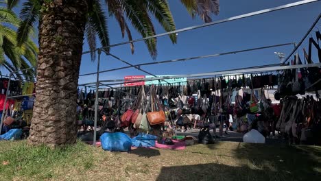 Street-street-street-peddler-selling-counterfeit-women's-handbags-and-replicas-for-tourists-under-a-palm-tree-on-a-sunny-summer-morning,-shot-traveling-to-the-left