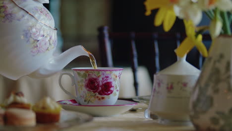 Teekanne-Wird-In-Teetasse-Auf-Einem-Schön-Gedeckten-Tisch-Mit-Blumen-Und-Gebäck-Gegossen