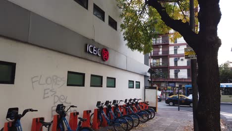 City-public-bicycles-parked-at-ICBC-in-bustling-avenue-traffic-of-buenos-aires-city,-flores-neighborhood,-argentine-capital,-Industrial-and-Commercial-Bank-of-China-establishing-shot