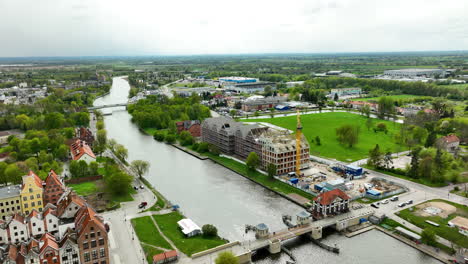 Aerial-view-of-a-riverside-urban-landscape-in-Elbląg,-with-construction-near-the-water's-edge-and-a-bridge-in-the-distance