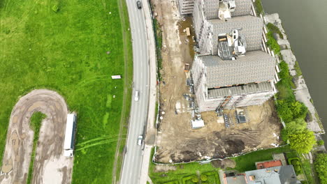 Aerial-construction-site-next-to-road-with-vehicles,-grass-field,-and-houses