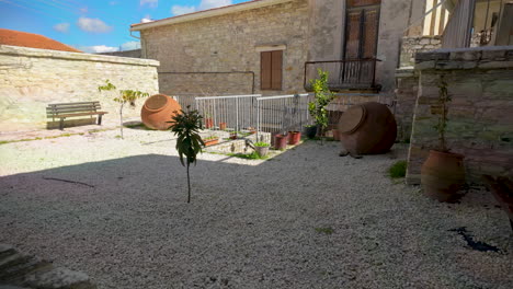 A-peaceful-stone-courtyard-featuring-gravel,-potted-plants,-and-large-clay-jars,-flanked-by-traditional-stone-buildings-in-Lefkara
