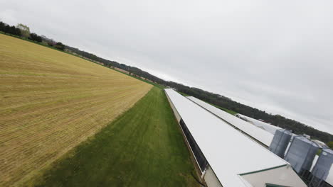 Fpv-drone-flight-over-farm-field-with-barn-for-animals-during-grey-sky-in-countryside-of-american-town