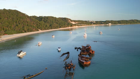 Boats-float-still-around-shipwreck-in-Tangalooma-Moreton-Island-forested-hills-landscape,-aerial-drone-flying-slow