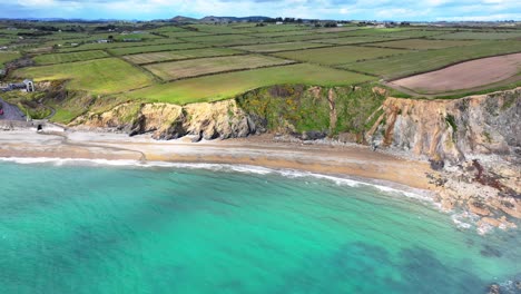 Drone-seascape-of-Killfaracey-beach-Waterford-Ireland-with-green-fields-sloping-down-to-cliffs-and-the-sea-and-emerald-clear-seas