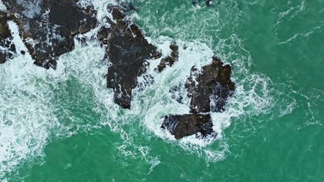 Drone-top-down-ascend-above-ocean-waves-pulsing-across-rocks-breaking-surface