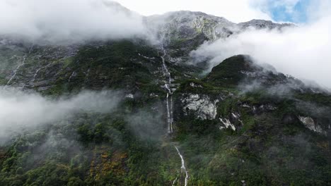 Incredible-thin-waterfall-descends-cascading-between-clouds-and-forest-in-Milford-Sound,-aerial-parallax
