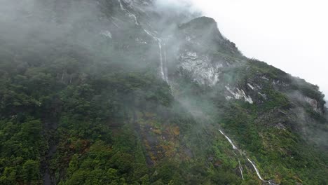 Misty-clouds-cover-steep-cliffs-revealing-cascading-thin-waterfall-through-forest-in-New-Zealand