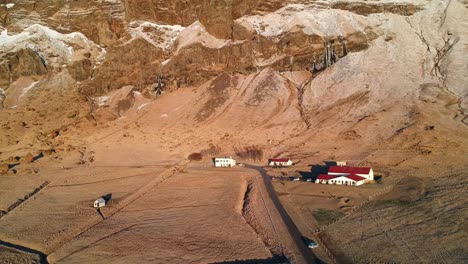 Aerial-shot-of-barren-landscape-in-south-coast-of-Iceland,-with-a-few-red-roof-houses-on-a-big-expanse-of-land-with-the-backdrop-of-tall-mountains-bathed-in-golden-light