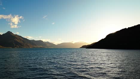 Stunning-sun-flare-retreats-behind-hillside-over-Lake-Wakatipu-with-grand-mountains