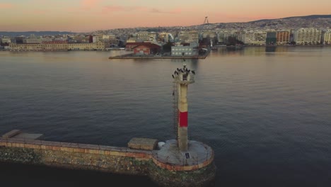 Lighthouse-During-Sunset-at-Thessaloniki-Waterfront:-Aerial-Drone-Showcases-Lighthouse-With-Birds,-Backdrop-of-the-City-Skyline---Captivating-Views-of-Greek-Urban-Coastal-Beauty-Θεσσαλονίκη,-Ελλάδ?