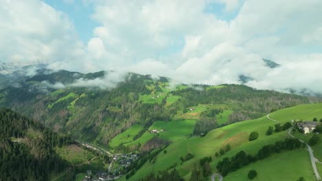 Un-Dron-Vuela-Rápido-Hacia-Un-Pequeño-Asentamiento-En-Una-Colina-Verde-Y-Exuberante-Cubierta-De-Nubes