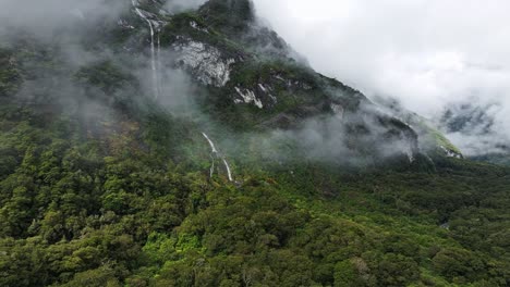 Sunlight-shines-on-dense-tropical-valley-with-clouds-wrapped-along-tall-waterfall-in-Milford-Sound
