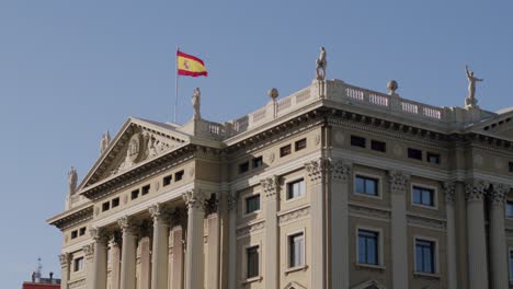 Bandera-Española-Ondeando-En-El-Gran-Edificio-De-Barcelona,-Fondo-De-Cielo-Azul,-Toma-Estacionaria