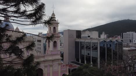 Aerial-crane-shot-climbs-pine-tree-in-plaza-near-Salta-Cathedral,-ARG